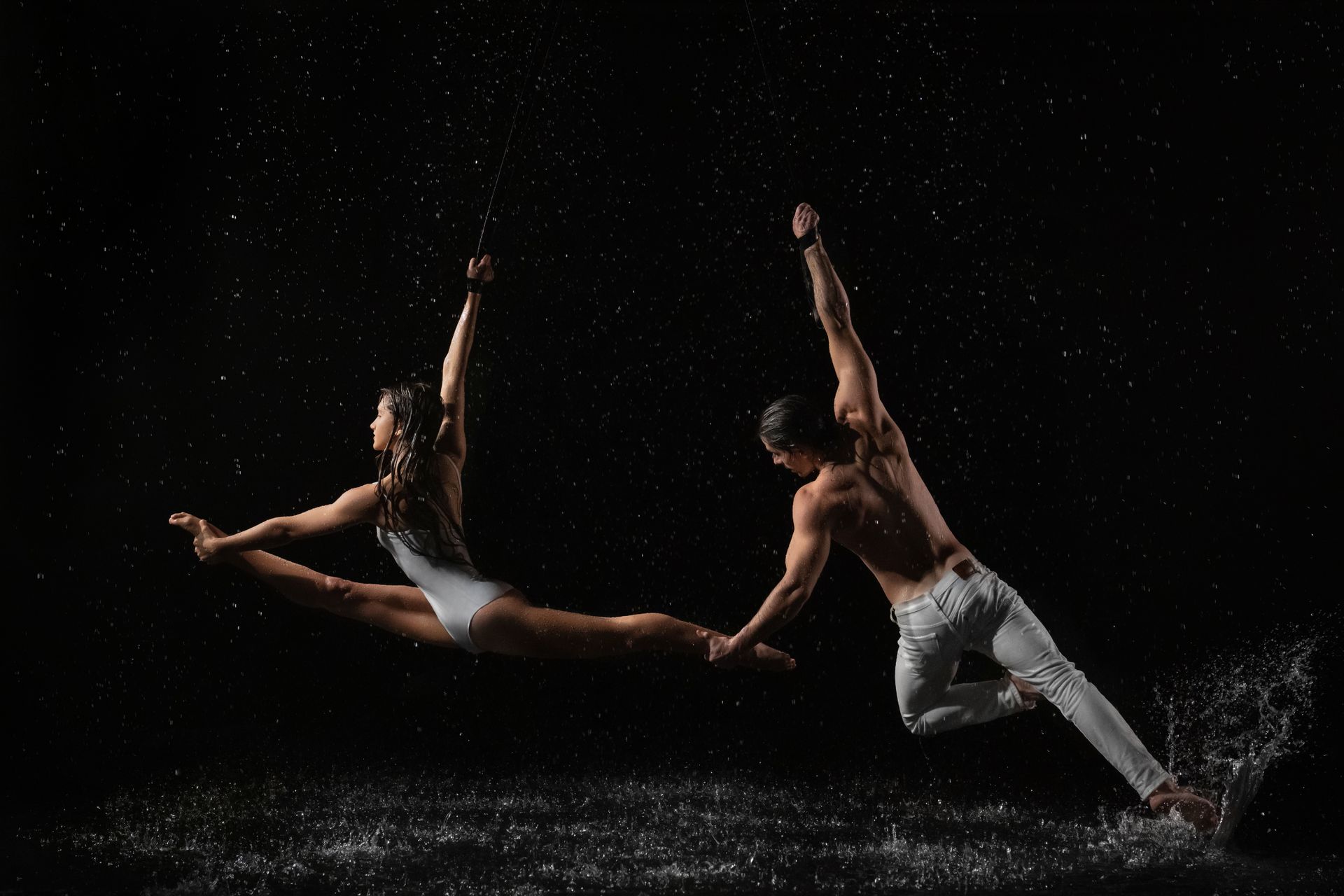 Male and female dancers perform a dynamic leap with water splashes, evoking movement and drama.