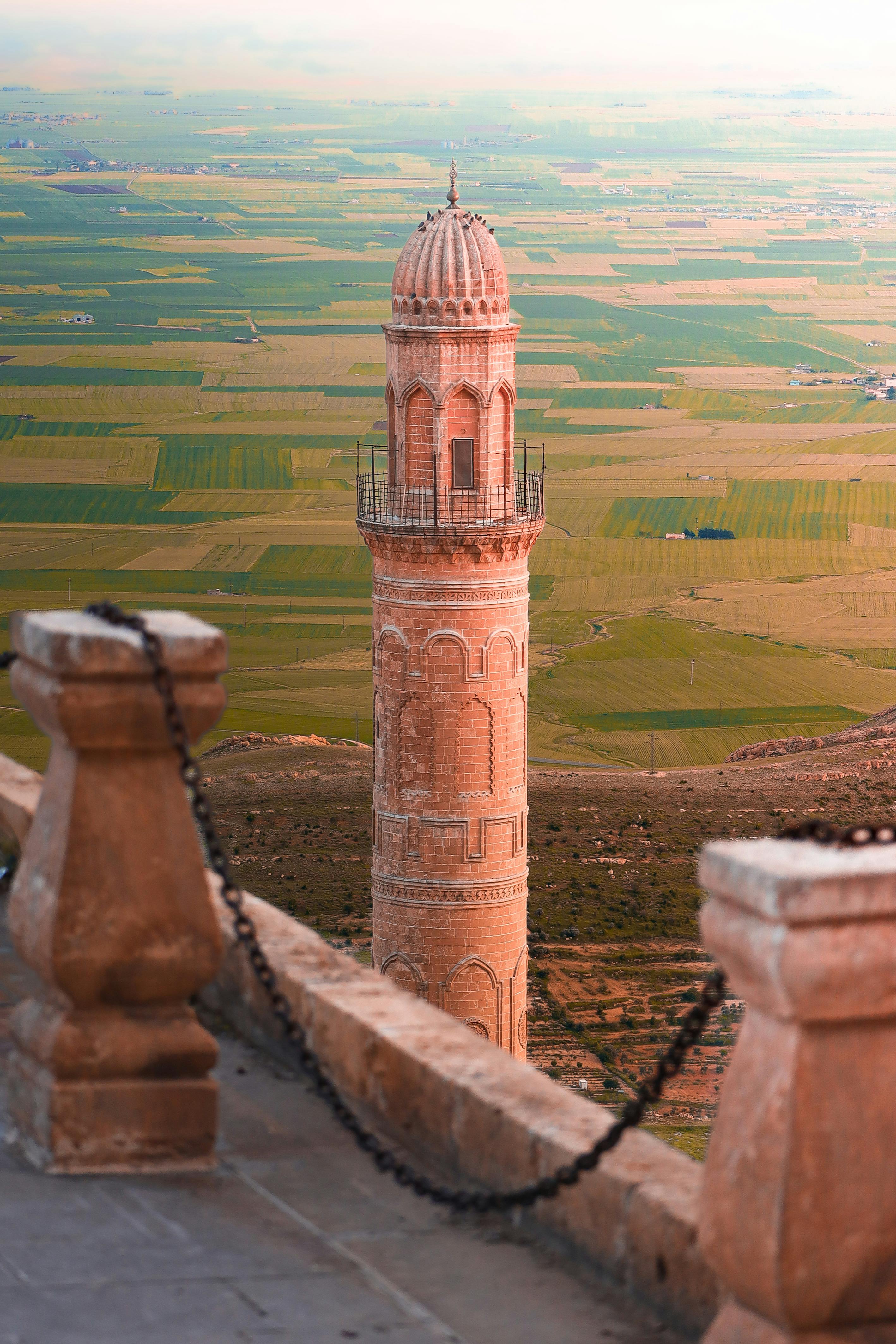 Ancient minaret in Mardin, Türkiye, against vast green plains, showcasing stunning Arabic architecture.