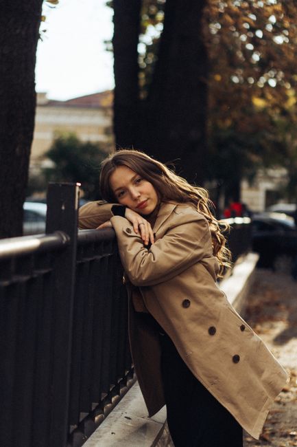 Stylish woman in trench coat leaning on a railing, capturing autumn vibes in the city.