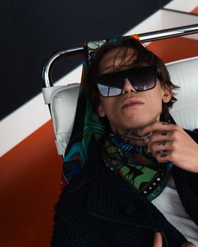 Portrait of a stylish man reclining with sunglasses and a colorful scarf, showcasing urban fashion elegance.