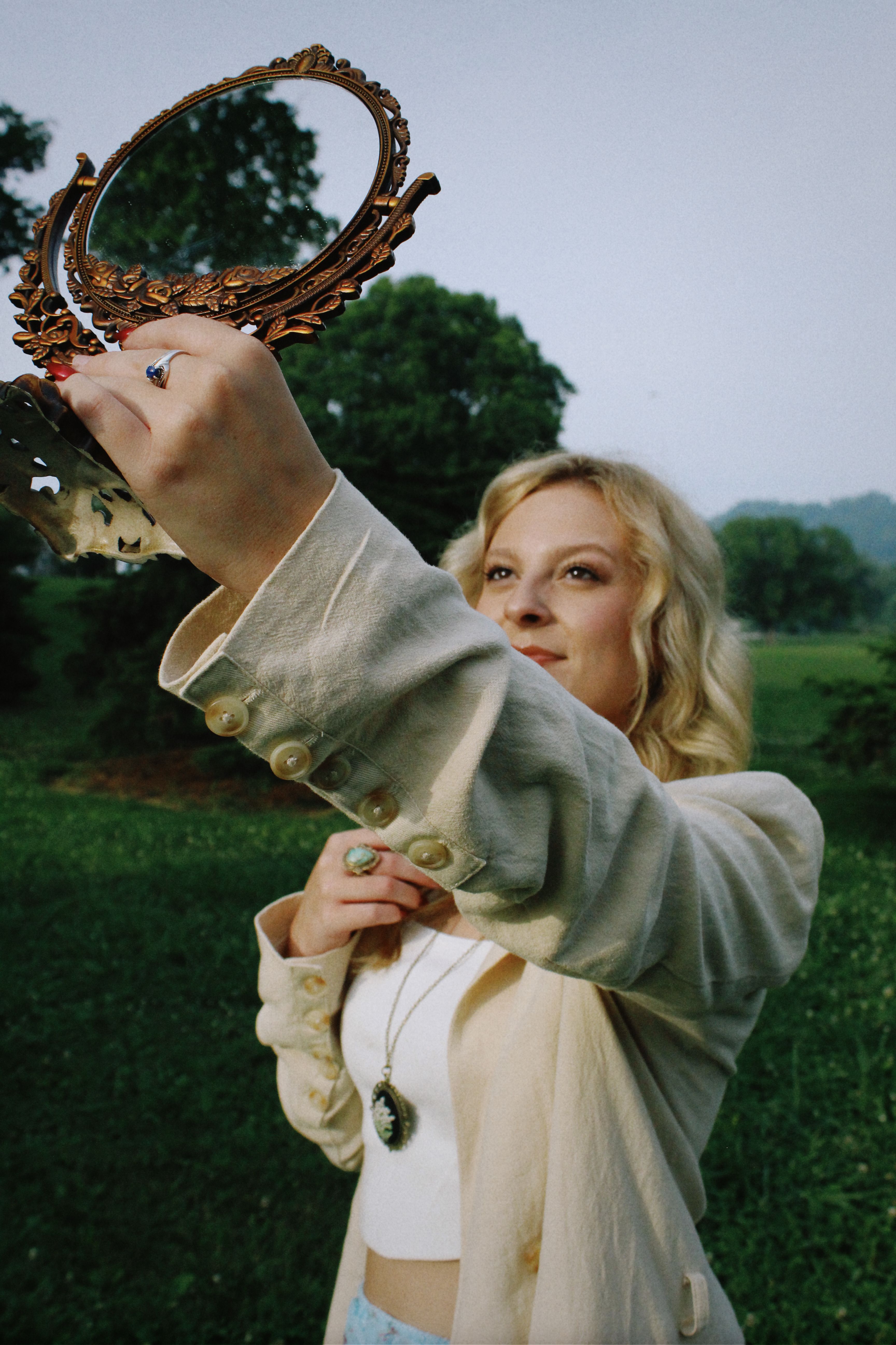 Blonde woman in countryside holding an ornate mirror, reflecting summer beauty.