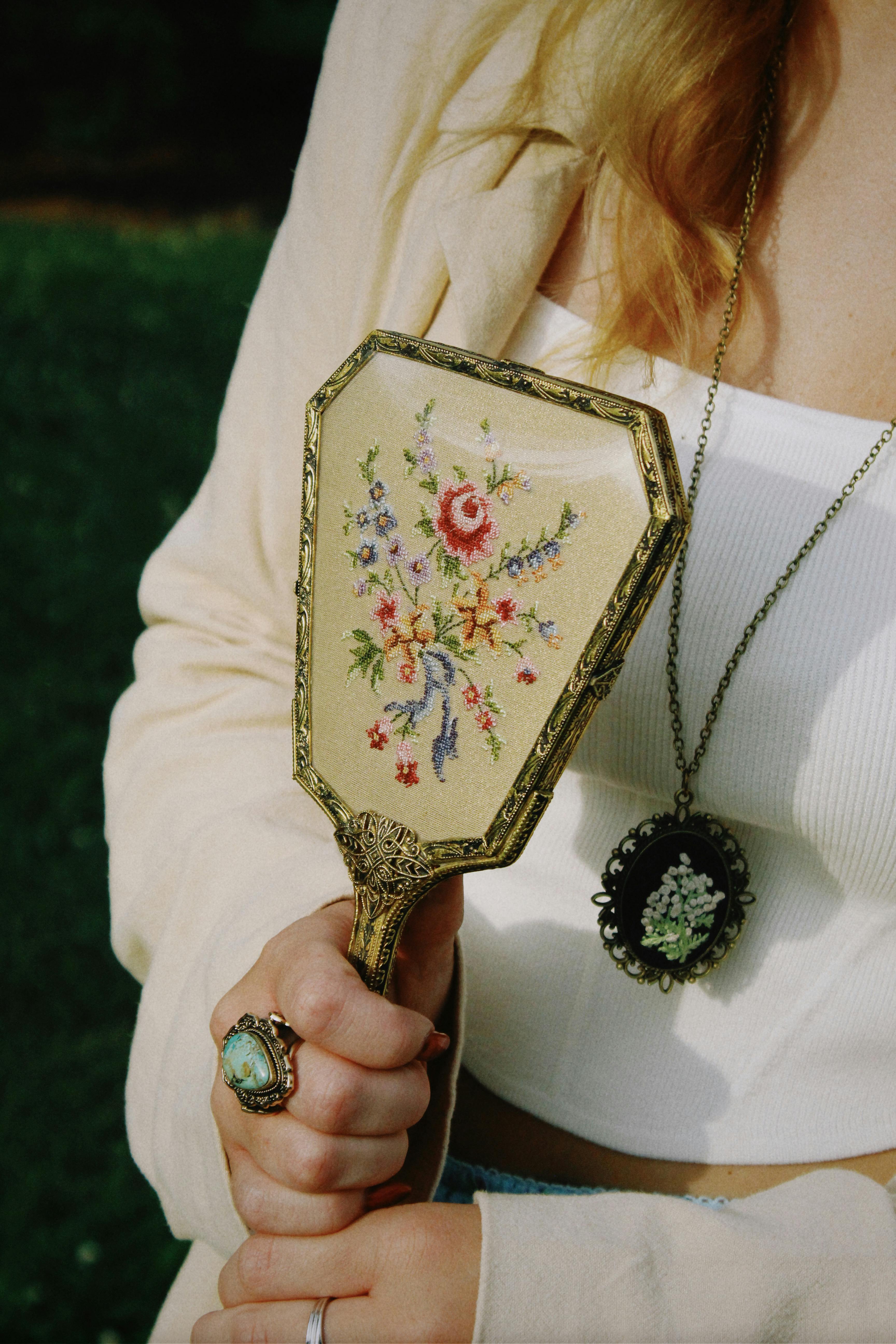A woman holding a vintage floral hand mirror with accessories in a natural setting.