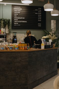 A warm café interior showing a barista working behind the counter with a menu board overhead.