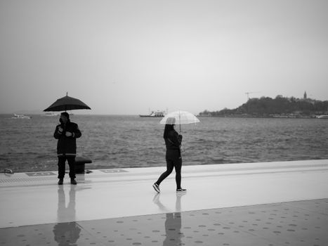 Two people with umbrellas walk along an Istanbul seaside promenade on a rainy day.