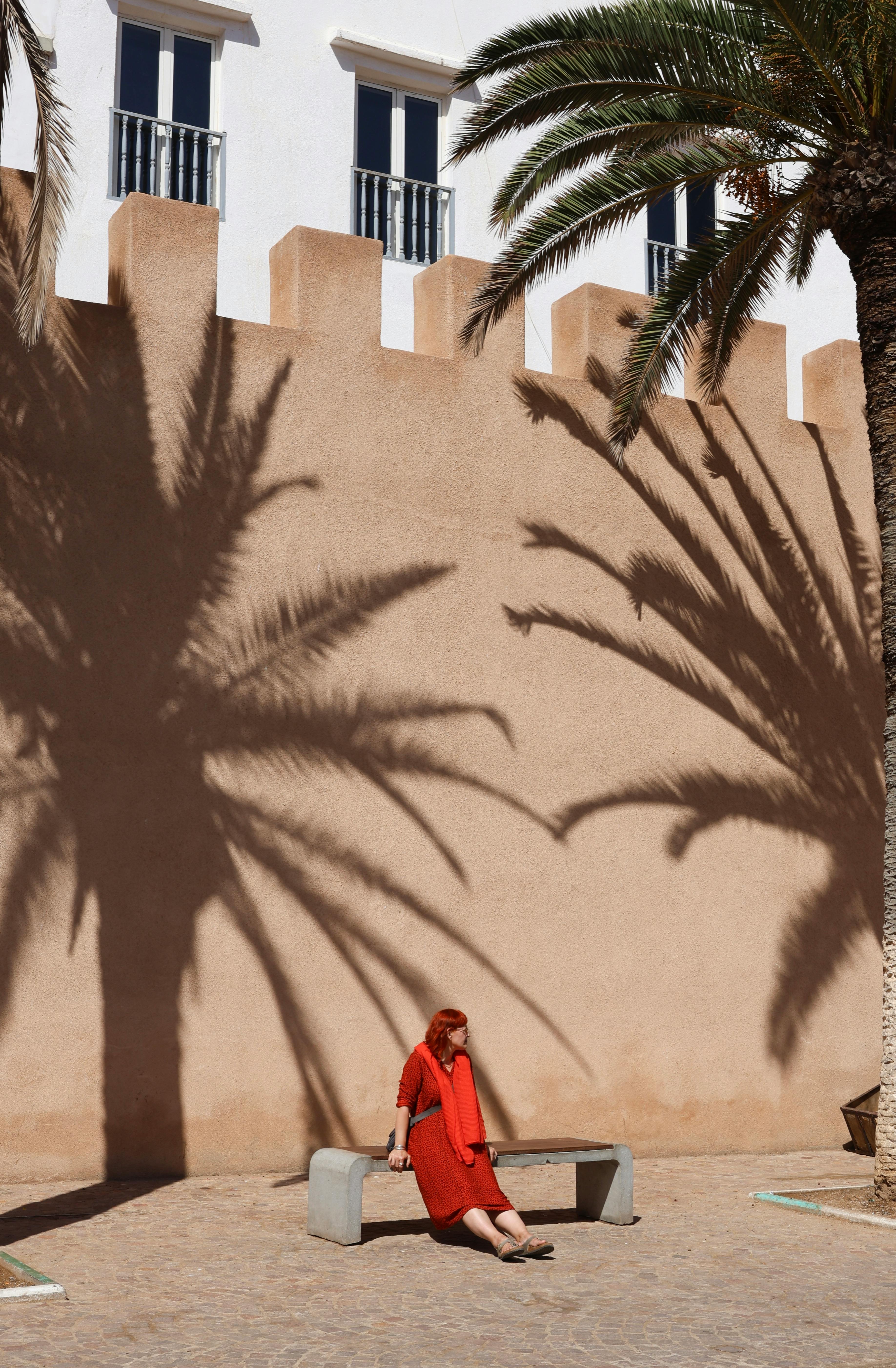 A woman in a red dress sits on a bench in urban sunlight, with palm tree shadows on the wall.