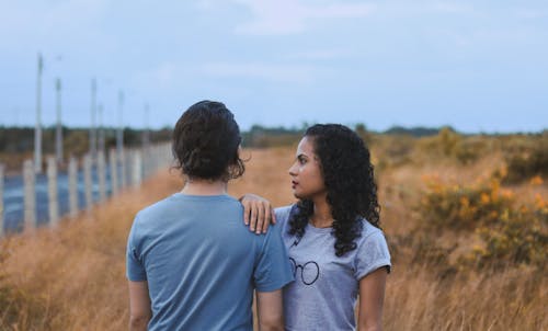 Young ethnic female with curly hair in casual wear touching shoulder of anonymous boyfriend while spending time together in dry field in countryside