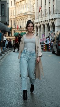 Stylish woman in trench coat walking on lively city street with historic architecture.