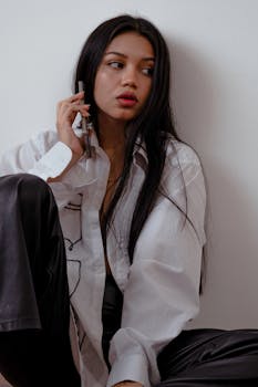 Fashionable woman in a white shirt sits indoors using a smartphone.