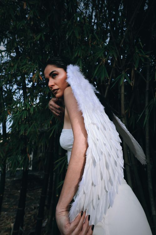 Free A woman with wings posing gracefully in a forest, wearing a white outfit and angelic wings. Stock Photo