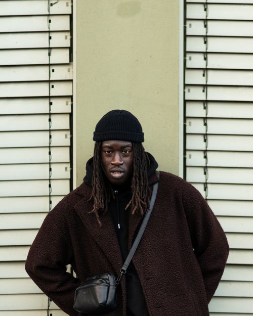 Free Fashionable man in a beanie and brown coat posing against a modern wall. Urban style and streetwear. Stock Photo