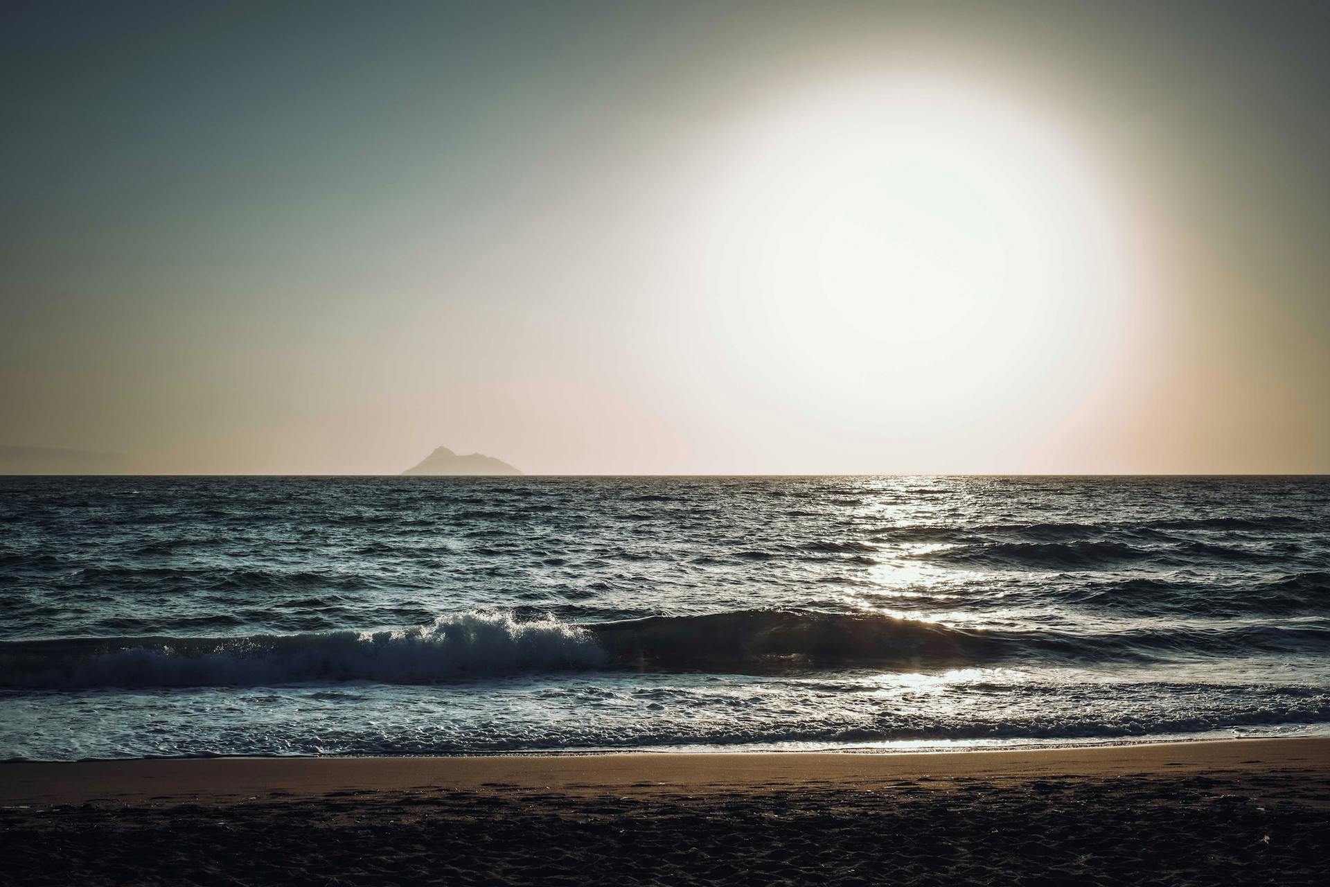 Tranquil beach scene at sunset with ocean waves and a distant island silhouette.