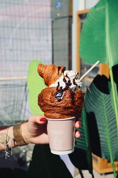 Hand holding a croissant topped with ice cream in a cup, outdoors.