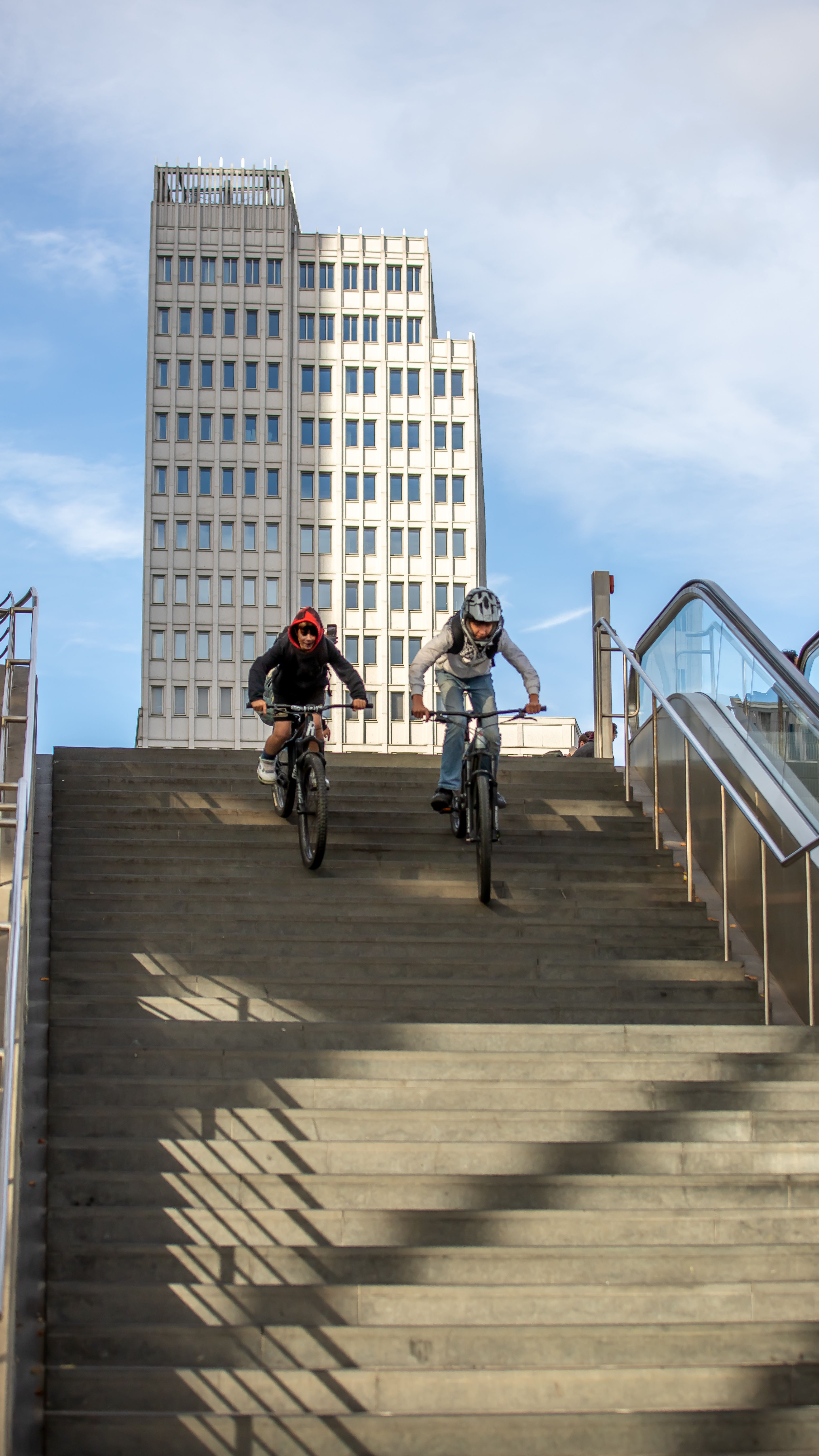 Two cyclists descending stairs with modern Berlin architecture in the background.