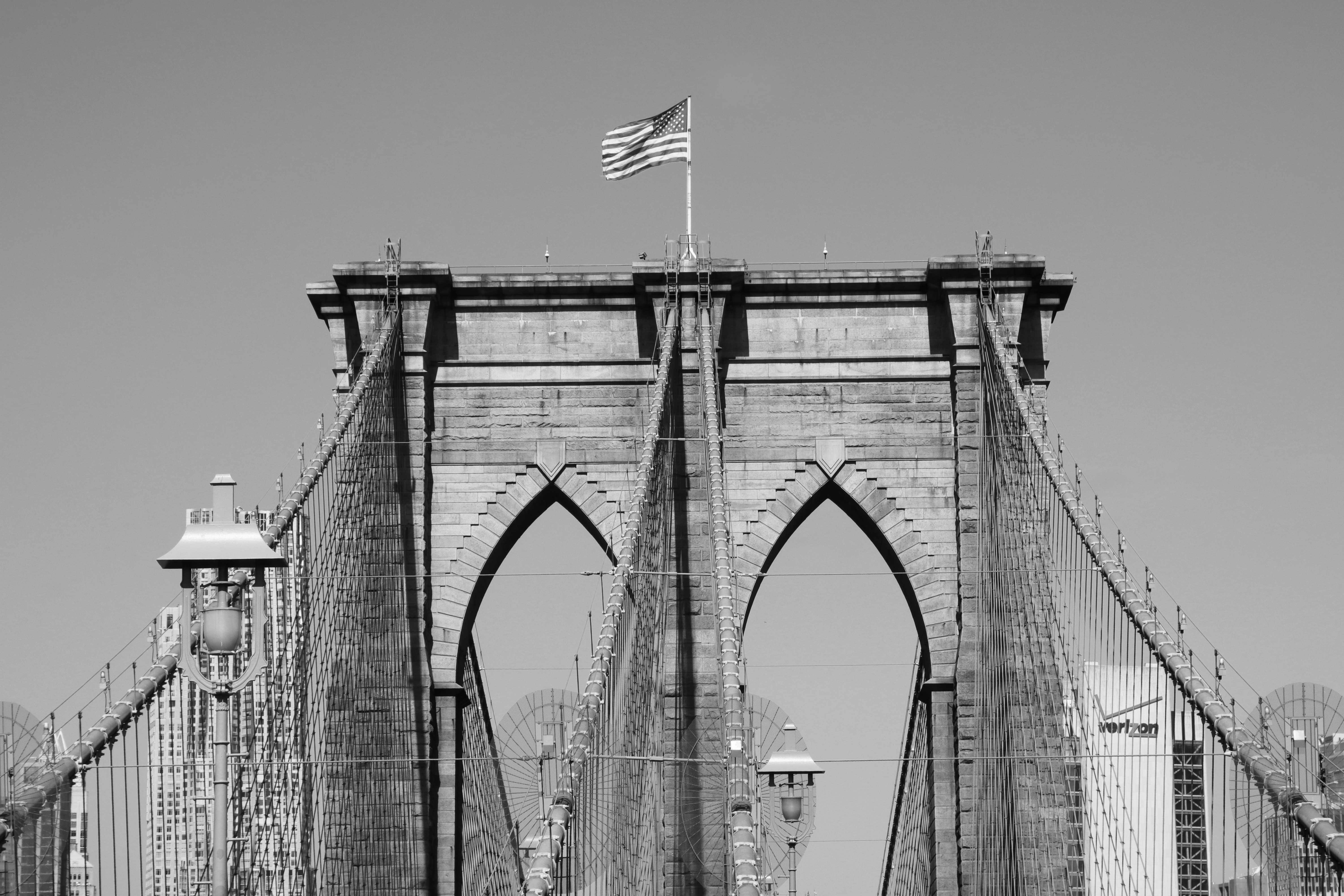 Black and white shot of Brooklyn Bridge with American flag, showcasing NYC architecture.