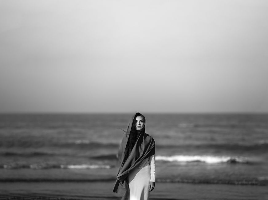A woman in a headscarf stands on a serene beach, gazing at the sea.