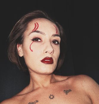 Portrait of a woman with bold makeup and tattoos, posing against a dark background.