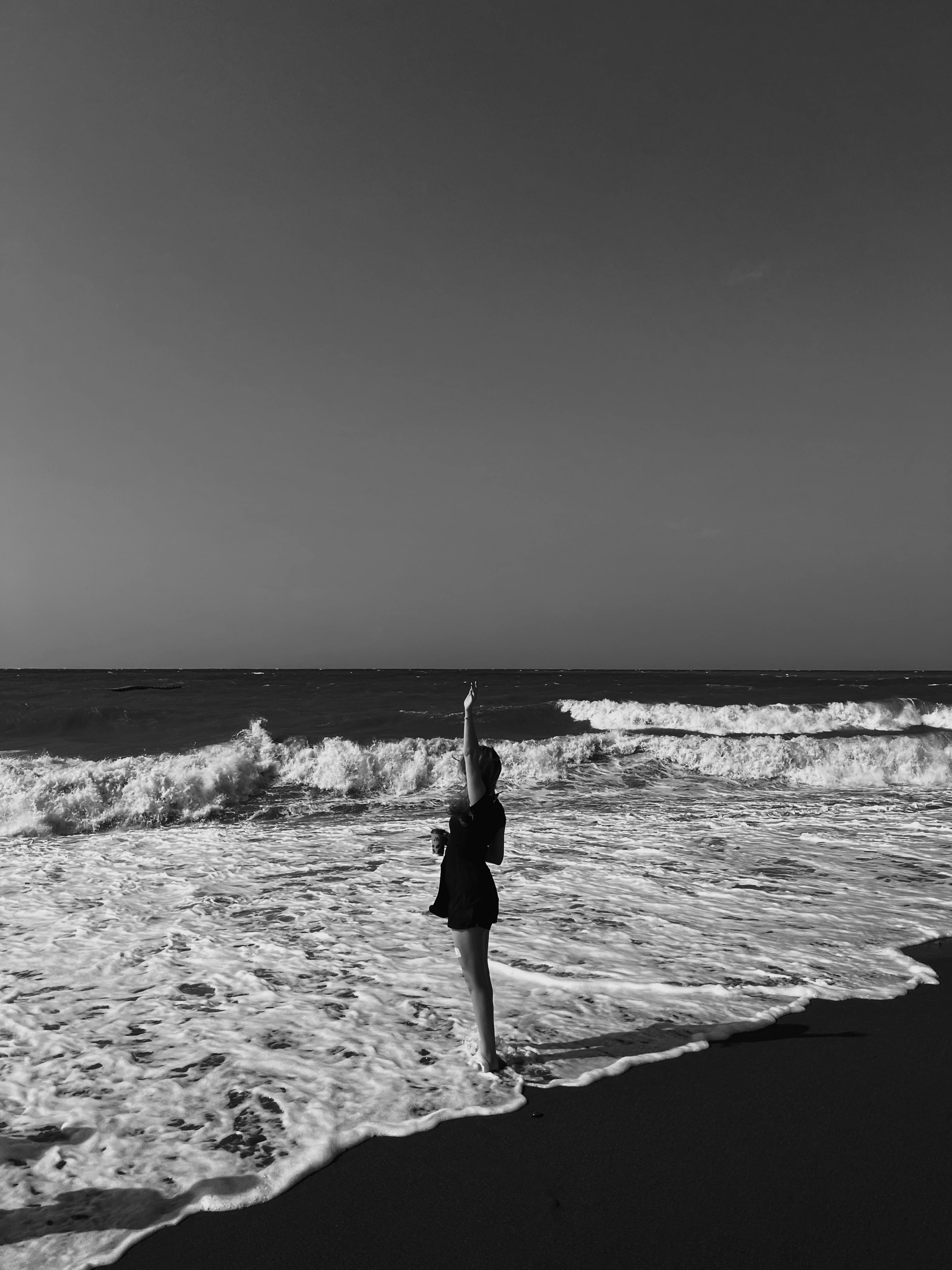 A woman stands by the sea with waves crashing, arm raised, in black and white.