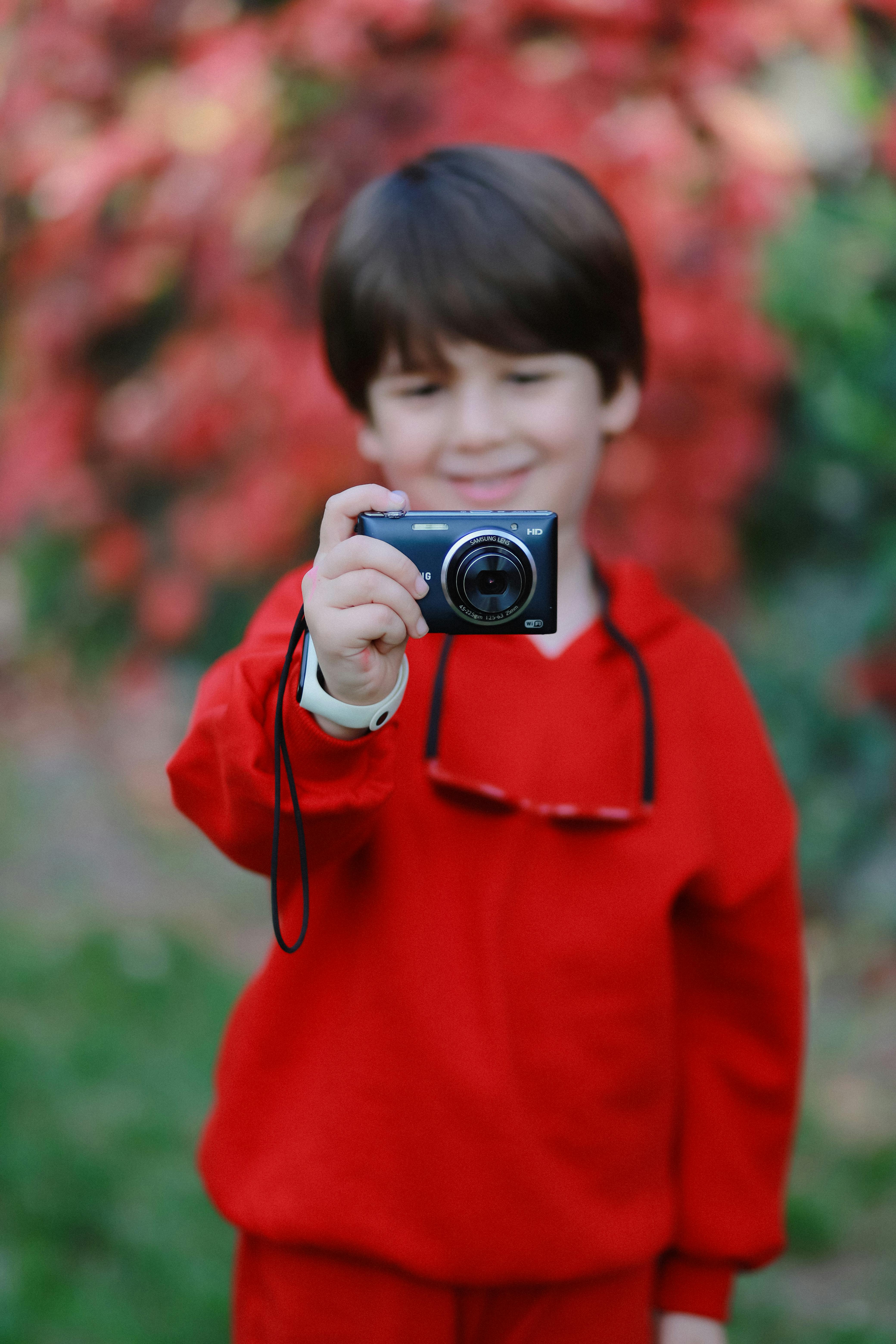 A cheerful boy in red clothes holding a camera outdoors amidst vibrant autumn colors.