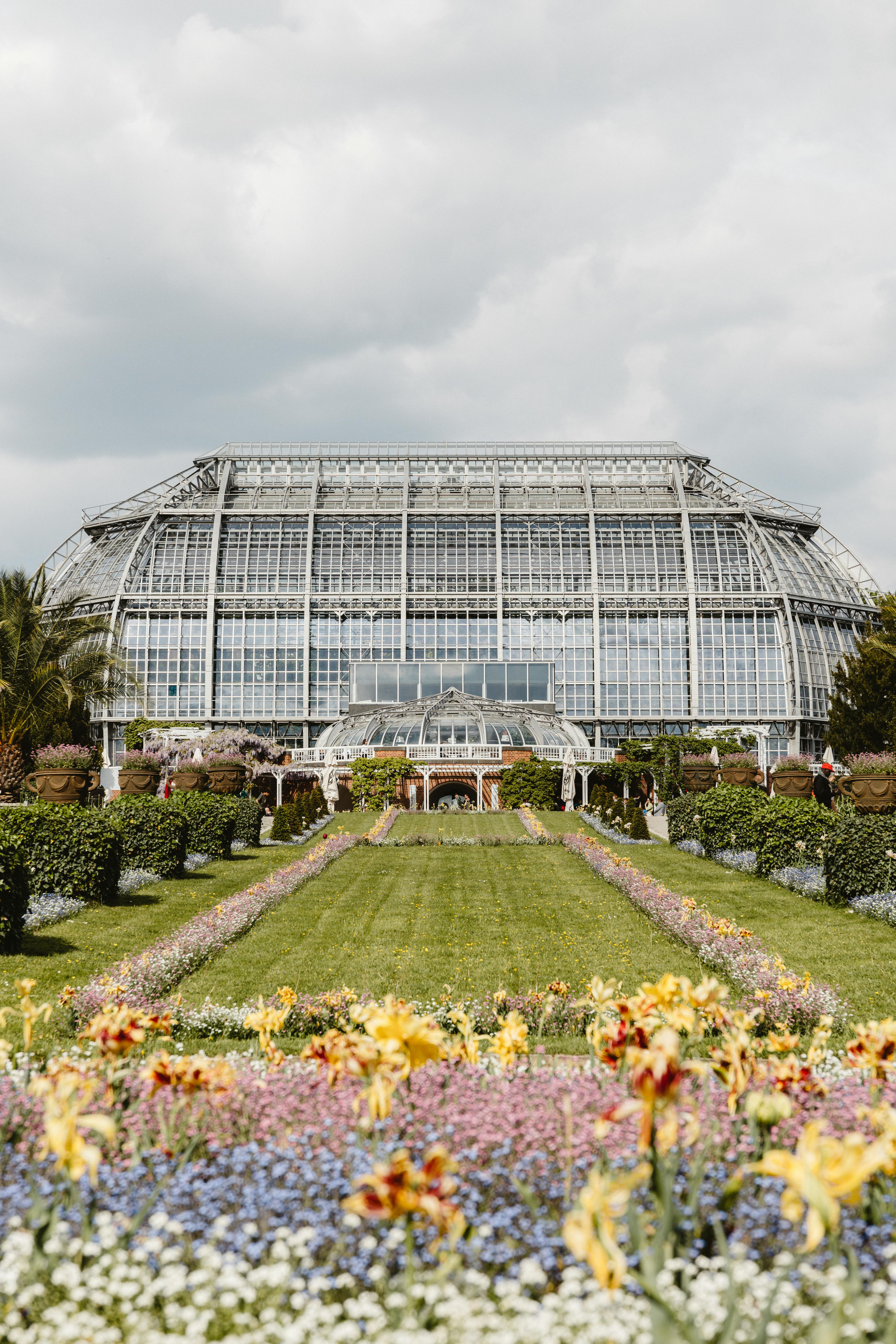Glasshouse in a vibrant Berlin garden under cloudy skies, showcasing floral splendor.