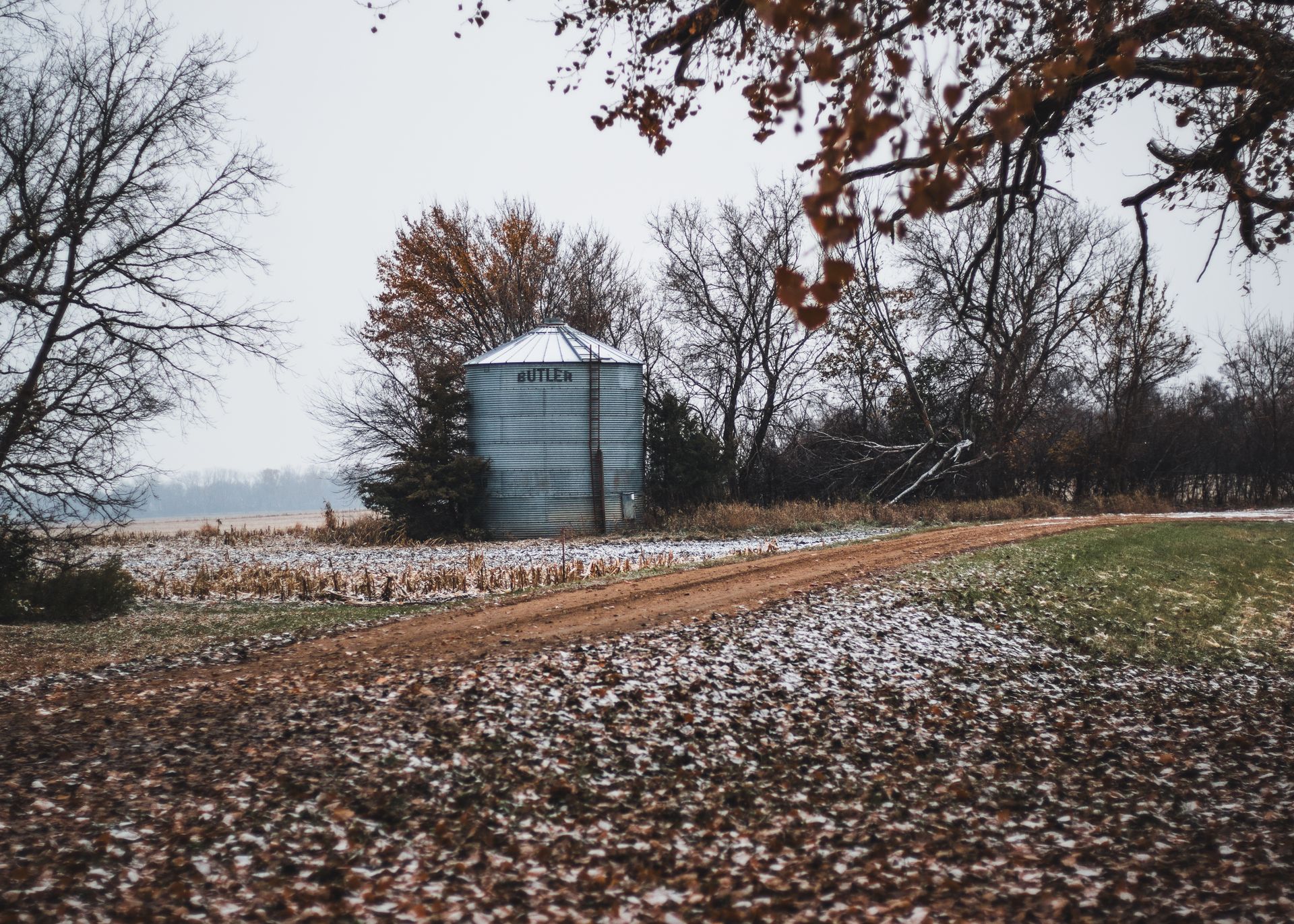 A serene winter scene featuring a rural dirt road and a grain bin surrounded by snowy fields and bare trees.