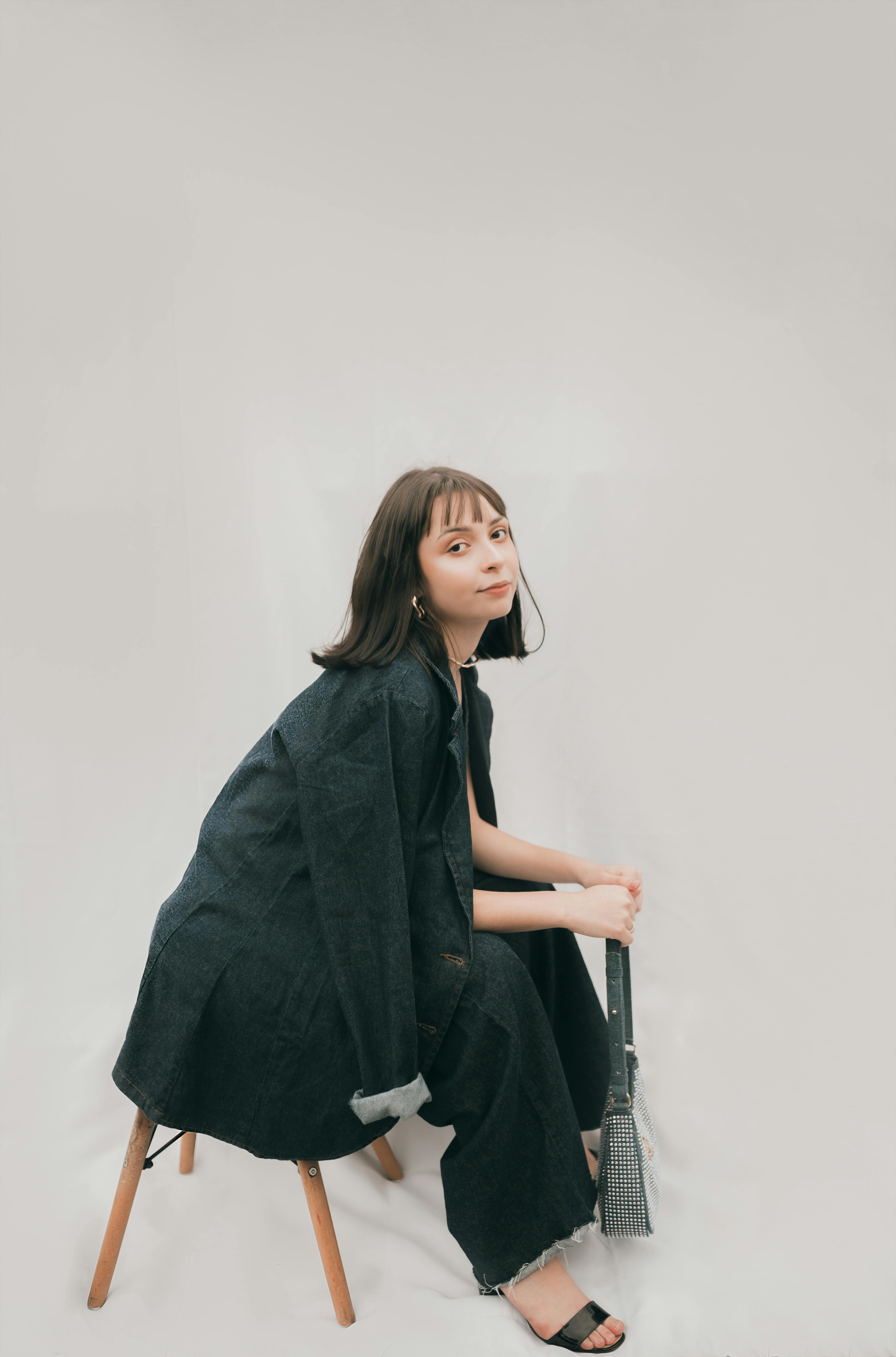 Chic woman sitting on a chair in a black jacket with a minimalist white background, exuding modern fashion.