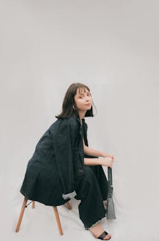 Chic woman sitting on a chair in a black jacket with a minimalist white background, exuding modern fashion.