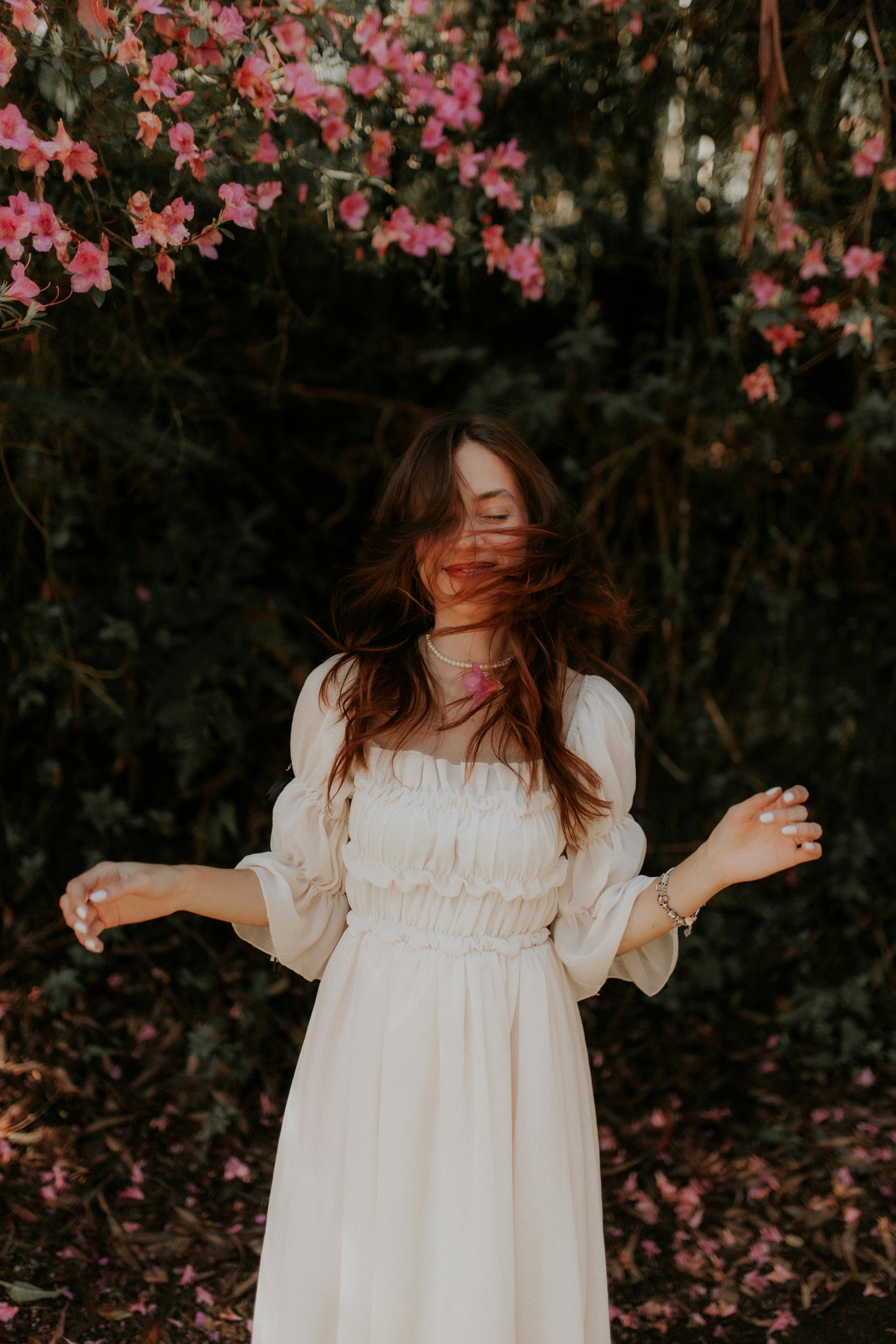Woman wearing white dress stands gracefully under vibrant pink blooming branches in a serene natural setting.