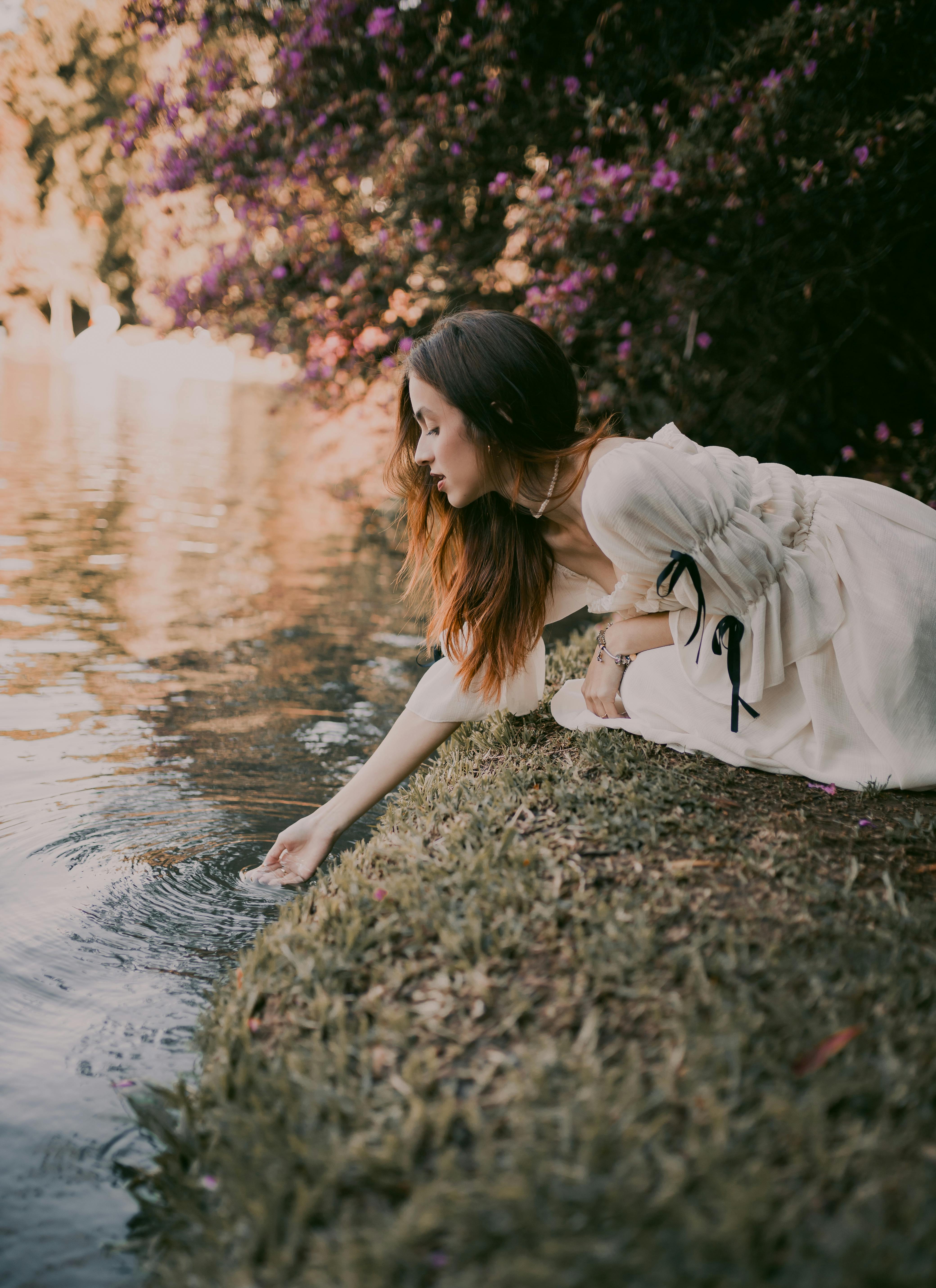 A woman in a vintage dress kneels by a riverside, immersed in nature's tranquility.