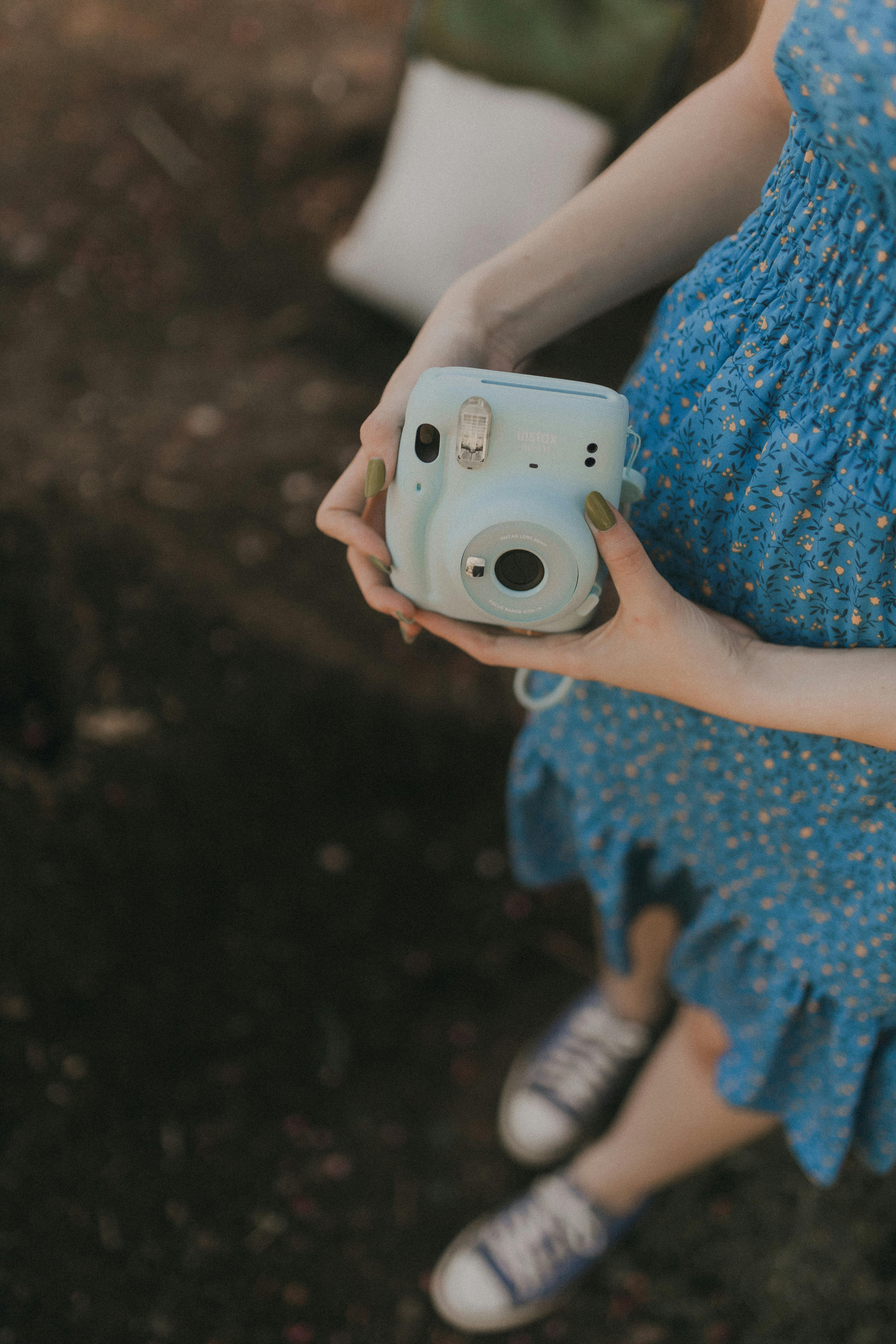 A woman in a blue floral dress holds an instant camera outdoors on a sunny day.