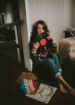 Woman sitting on the floor holding a vinyl record, surrounded by album covers in a cozy room.