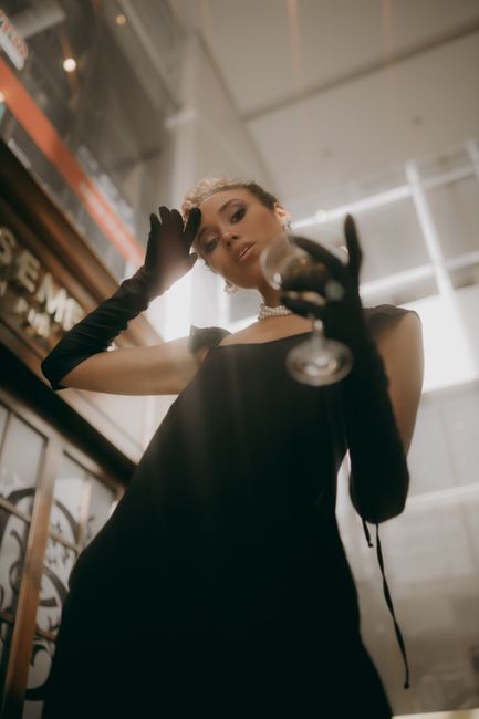 A fashionable woman in a black dress holds a glass elegantly indoors.