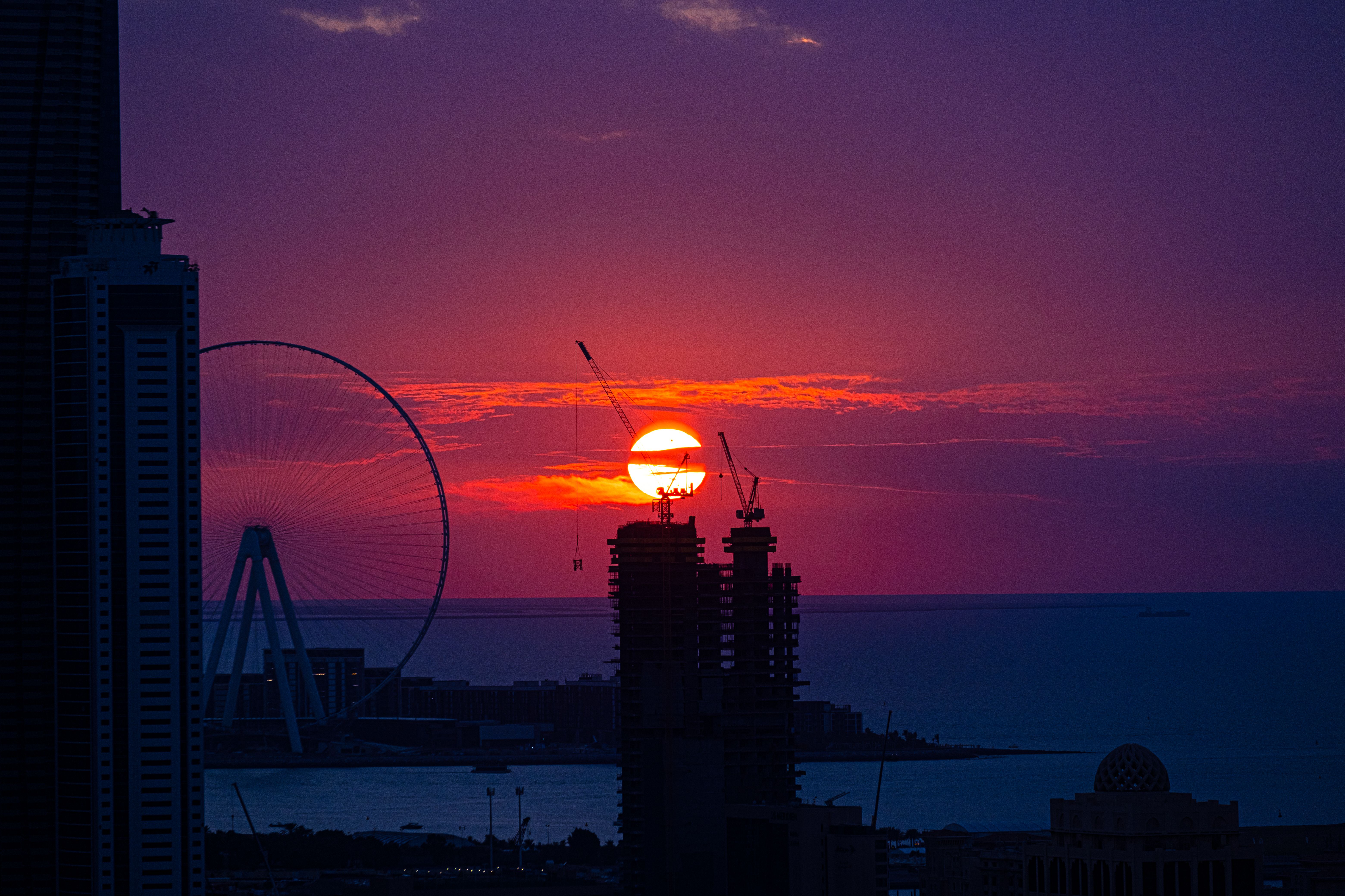 A breathtaking sunset over a city with a silhouetted Ferris wheel and construction cranes.