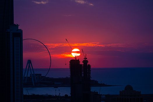 A breathtaking sunset over a city with a silhouetted Ferris wheel and construction cranes.