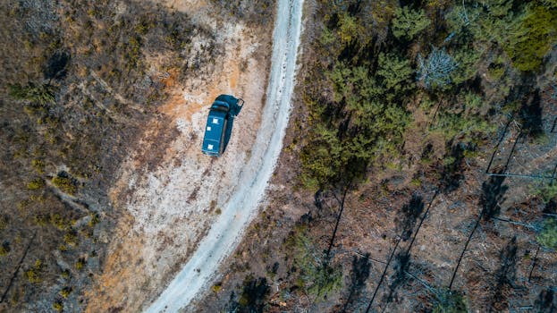 Bird's eye view of a vehicle driving through a rural forested dirt road during the day.