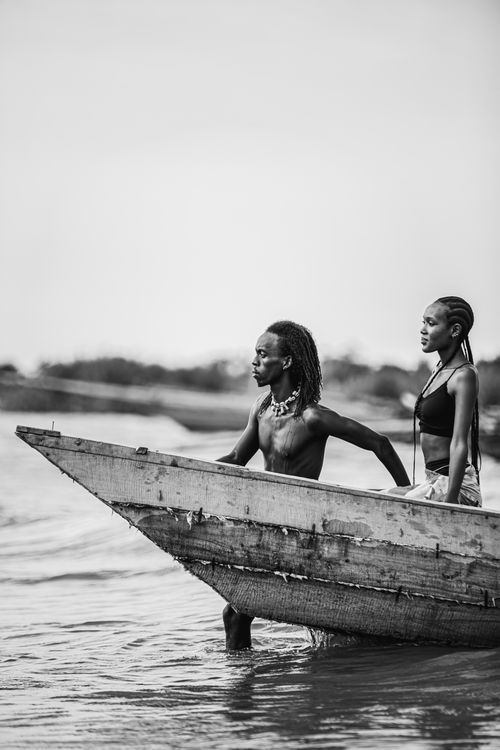 Free A couple stands in a wooden boat by a lakeshore, captured in black and white. Stock Photo