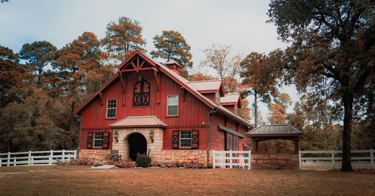 Picturesque red barn surrounded by lush autumn trees and a white fence, capturing serene countryside life.