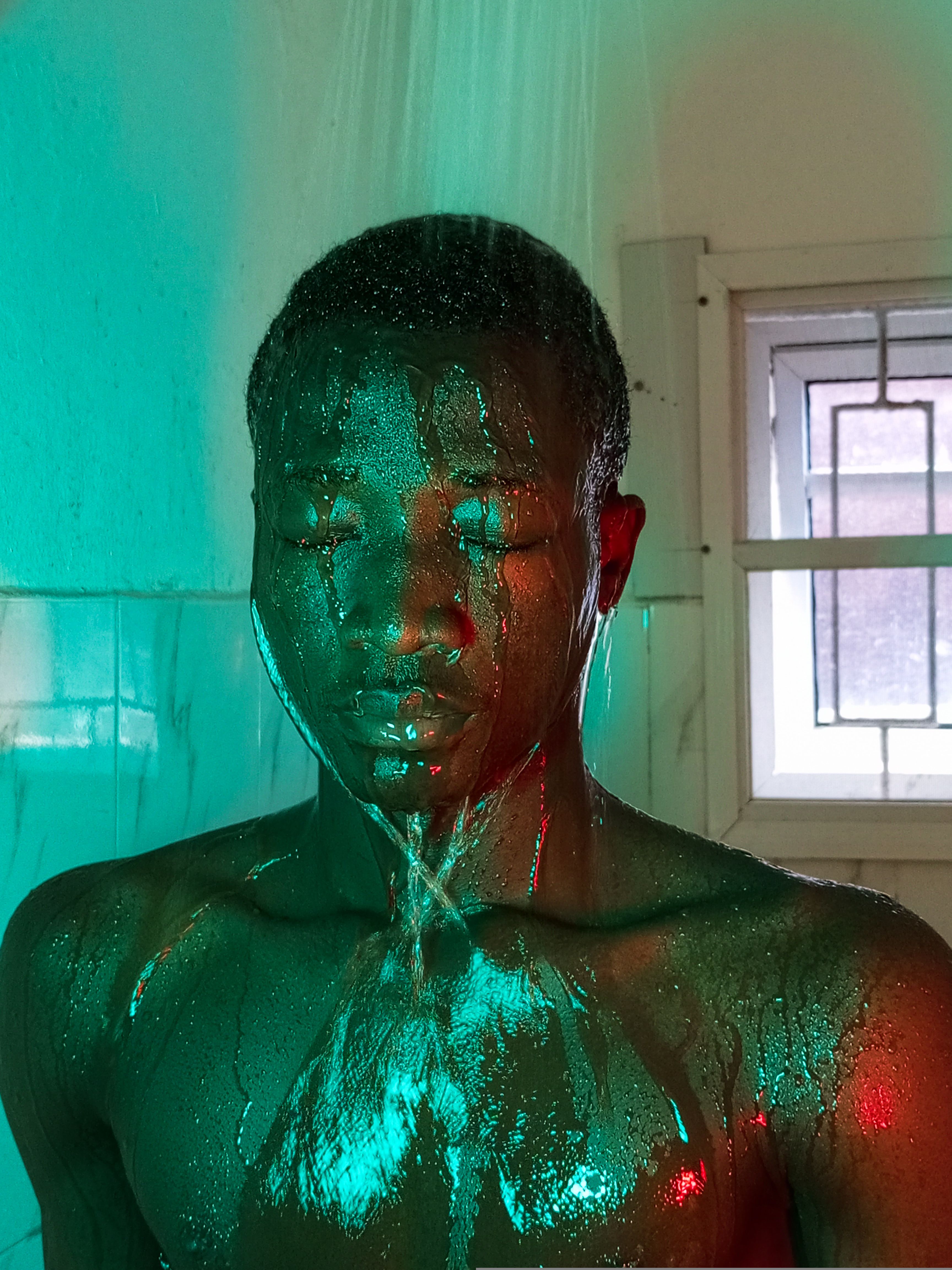 A young black man with eyes closed in a shower, colorful lighting creating a serene atmosphere.