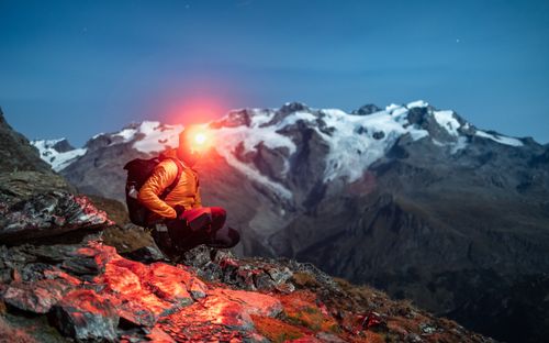 Free Hiker wearing headlamp kneels on rocky terrain with snowy mountains in the background at night. Stock Photo