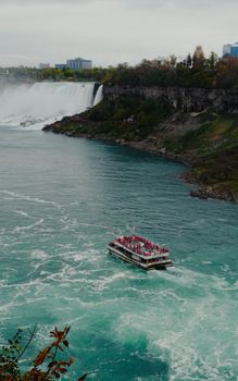 Boat cruising by a grand waterfall surrounded by lush cliffs under an overcast sky.