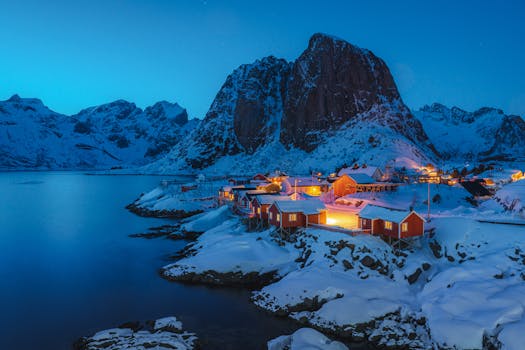 Beautiful aerial view of a snow-covered Norwegian village by the coast at night, surrounded by mountains.