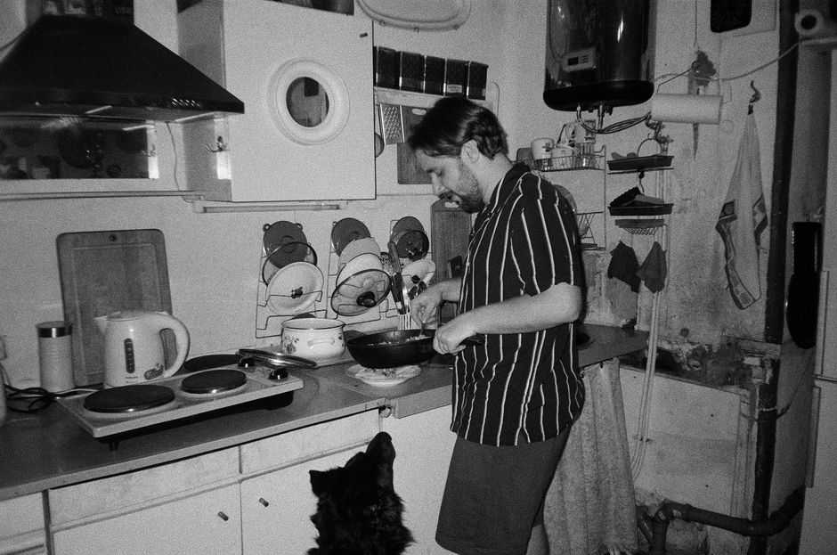 A man with short hair cooks in an old-style kitchen while a dog watches.