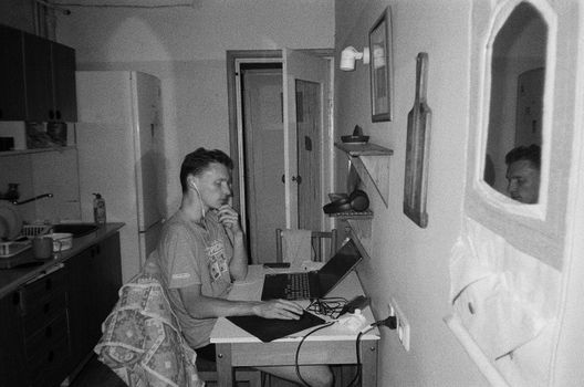 Black and white photo of a man working on a laptop in a kitchen setting, wearing earphones.