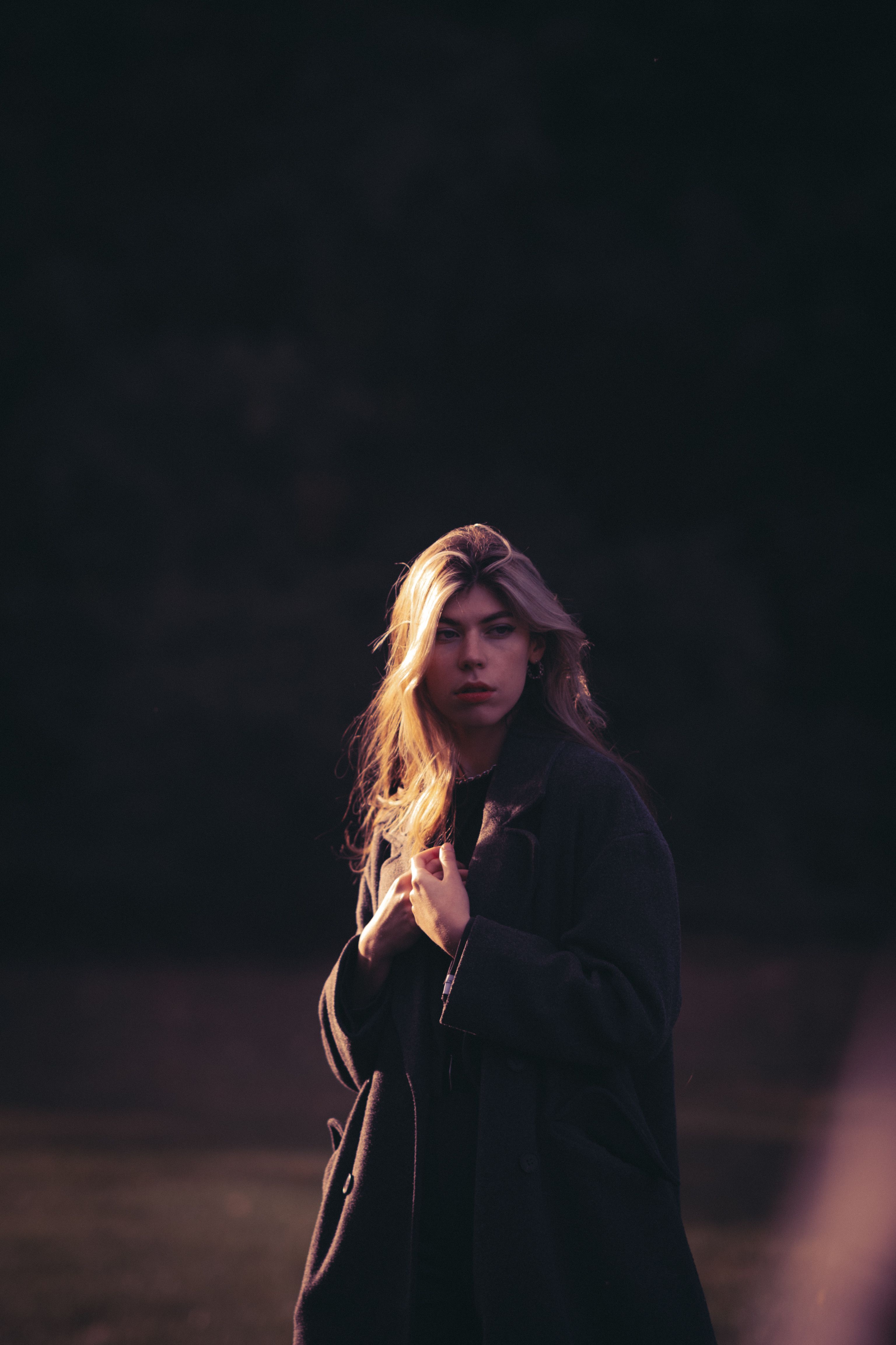 A moody portrait of a woman in a dark coat, captured outdoors with dramatic lighting.