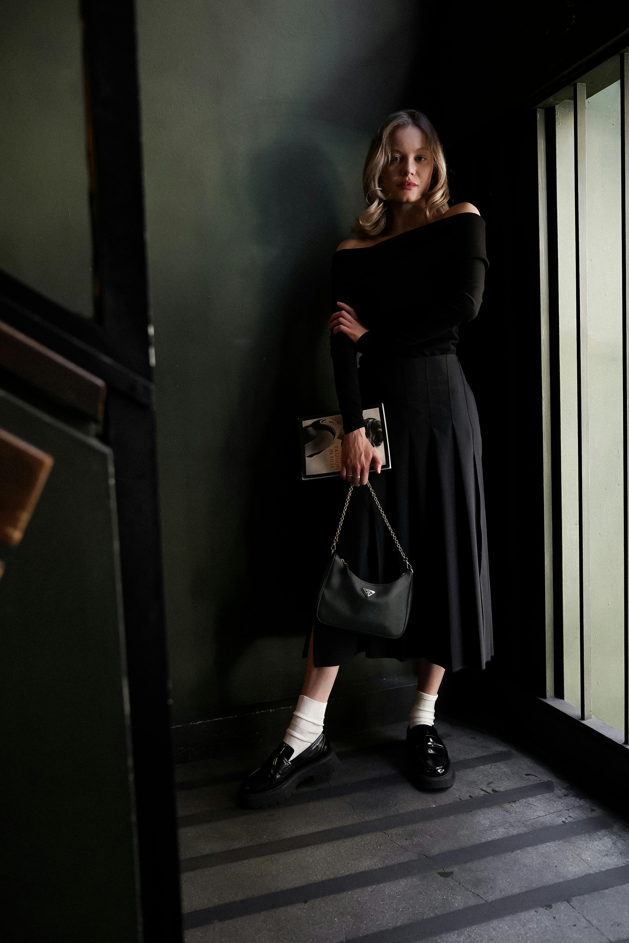 Elegant woman posing in chic black ensemble with purse and book indoors.