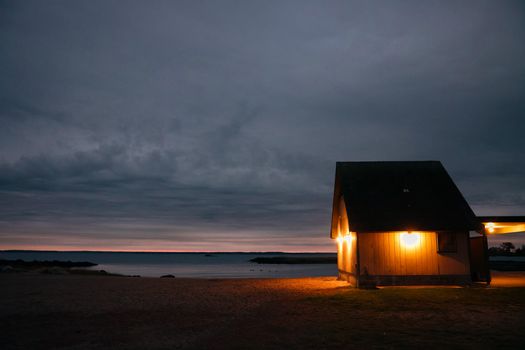 A warm-lit beach hut facing the calm sea at twilight, under a moody sky.