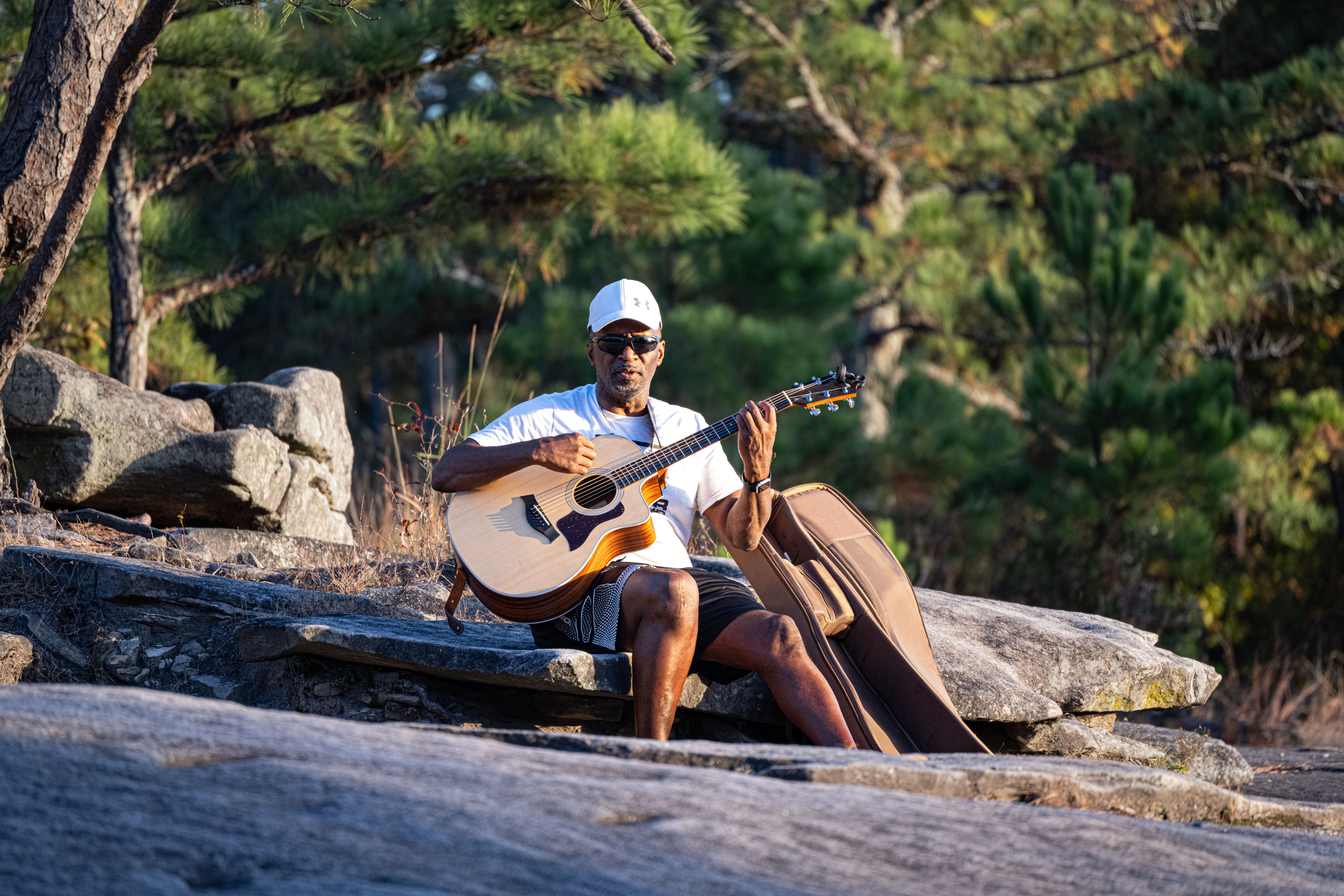 Musician in casual attire playing an acoustic guitar in a natural outdoor setting.