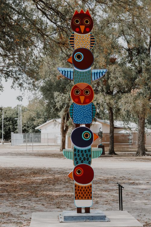 Free Vibrant owl-themed totem sculpture in an outdoor park setting with trees. Stock Photo