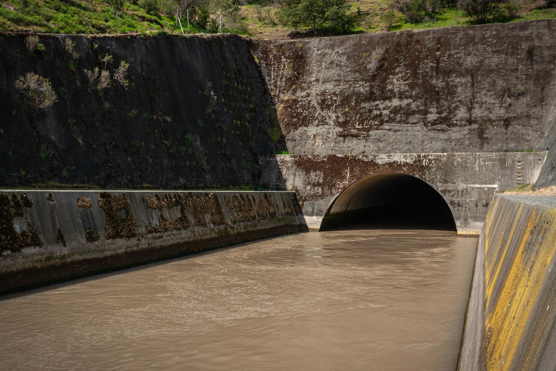 A large concrete storm drain with murky water flowing into a tunnel, surrounded by mossy walls.