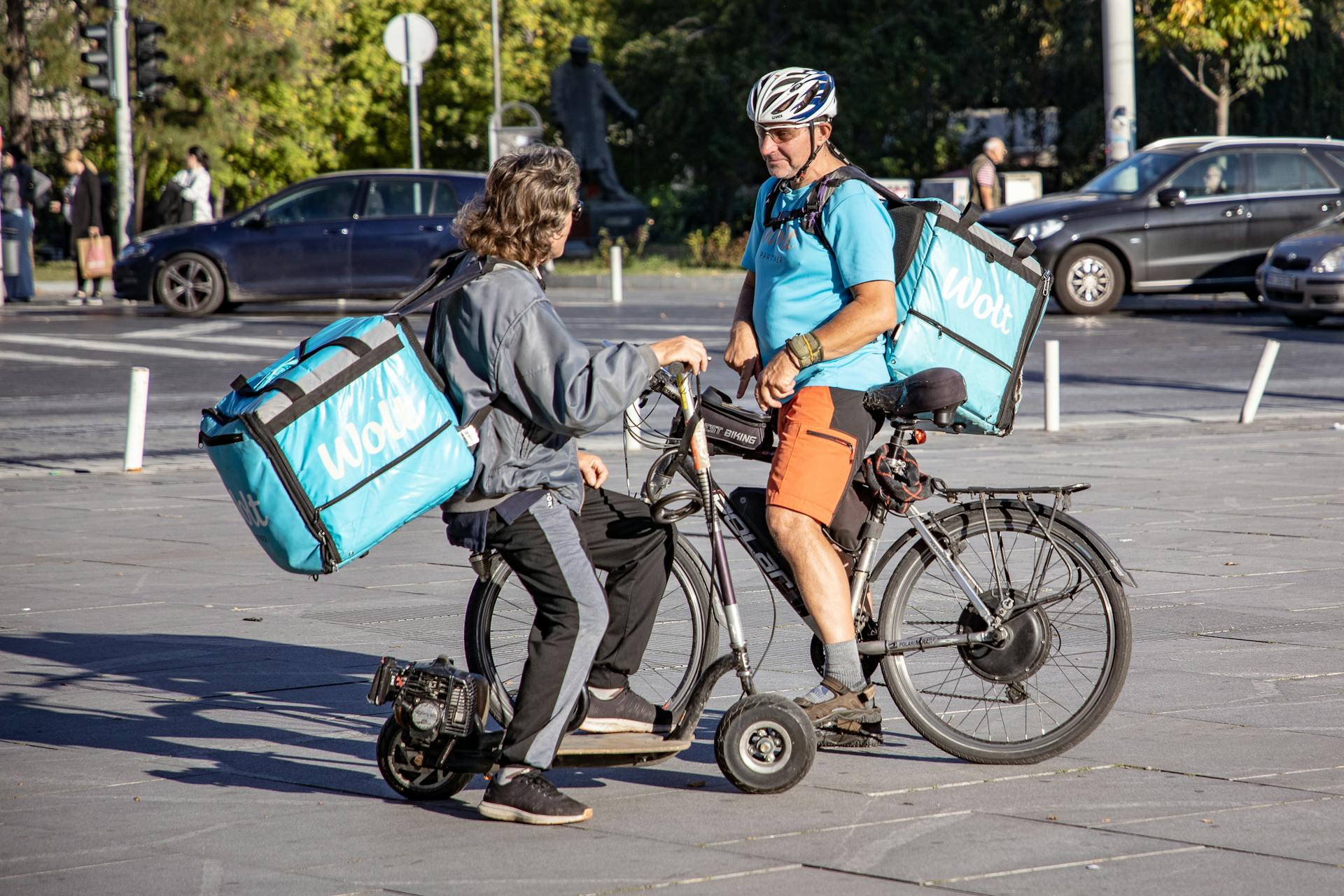 Two delivery couriers on bikes having a discussion on a city street.