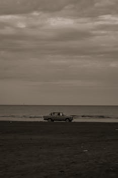 A vintage car parked on a Baku beach with a serene sea backdrop, captured in sepia tones.